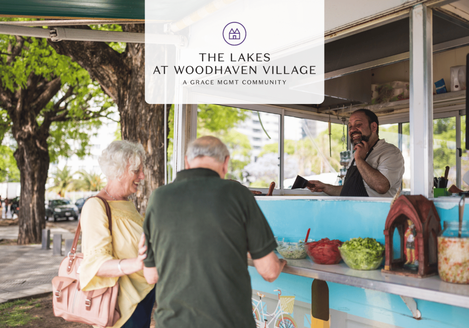 Outdoor food cart in a park with a smiling vendor serving two seniors; overlay reads 'The Lakes at Woodhaven Village' on a white banner with a logo.