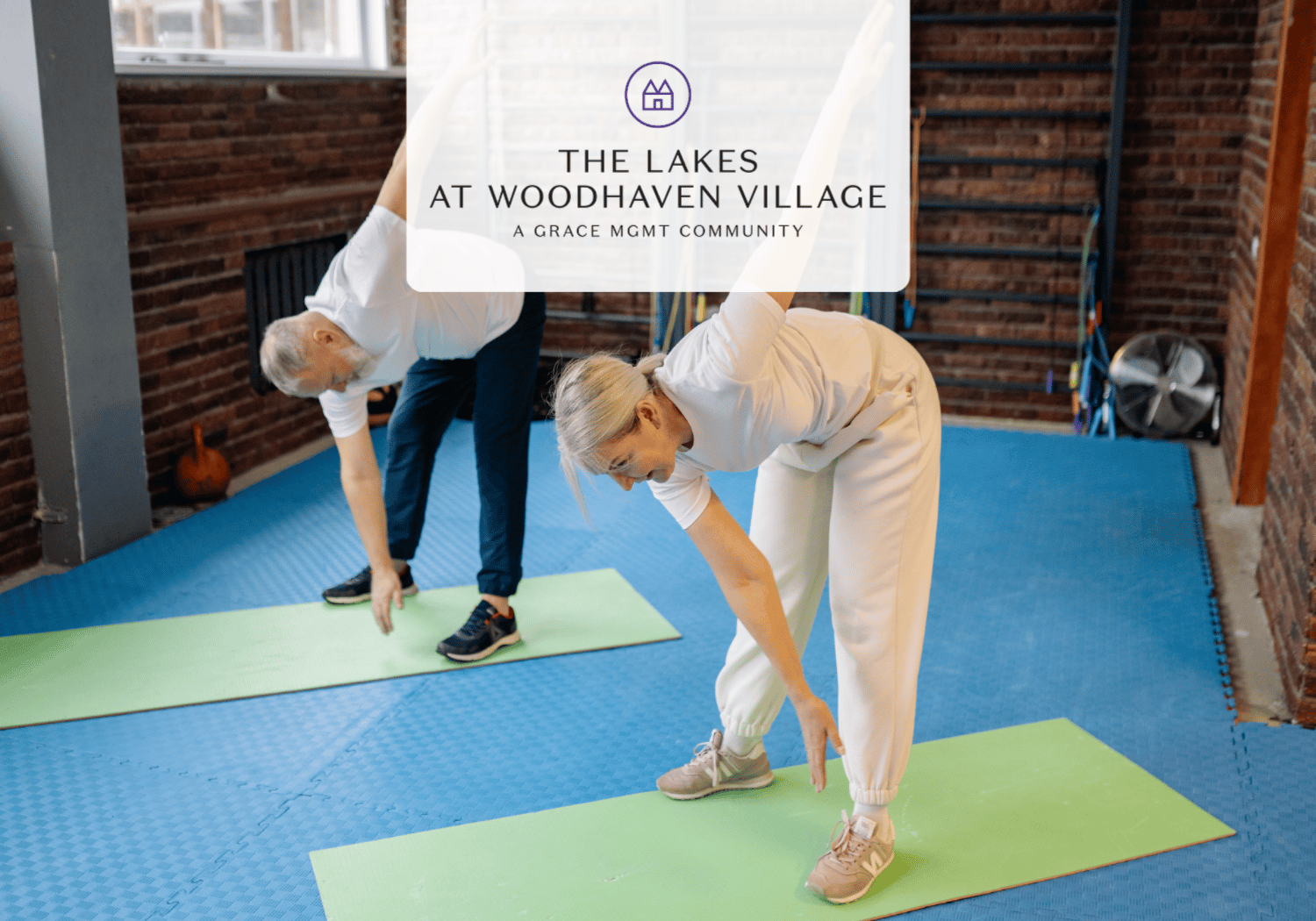 Two people in white outfits bend over on green exercise mats in a brick-walled studio, with a translucent overlay showing a logo and text 'The Lakes at Woodhaven Village' in the center.