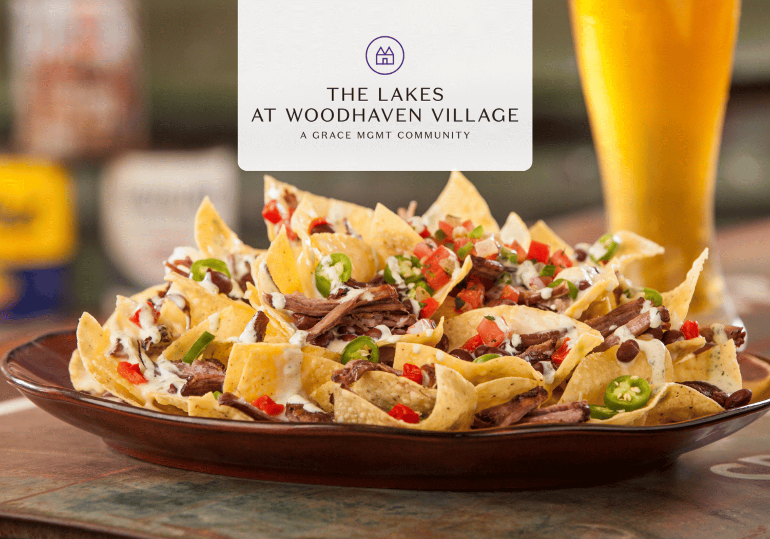 Plate of loaded nachos with beef, cheese, jalapeños, peppers, and sour cream; blurred background and a white logo reading The Lakes at Woodhaven Village above.