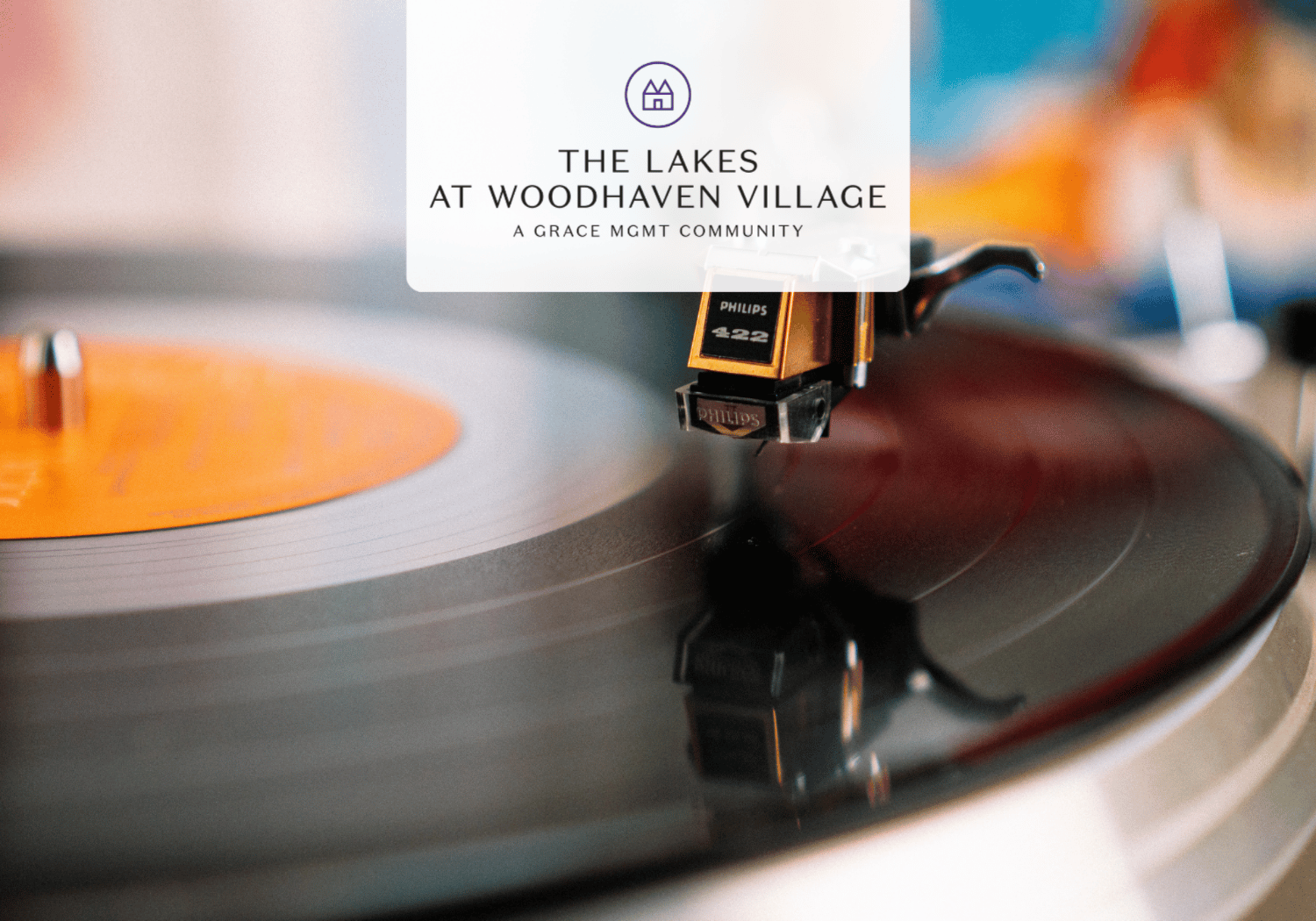 Close-up of a vinyl record spinning on a turntable with an orange label and a white rounded badge displaying 'The Lakes at Woodhaven Village' (advertising banner).