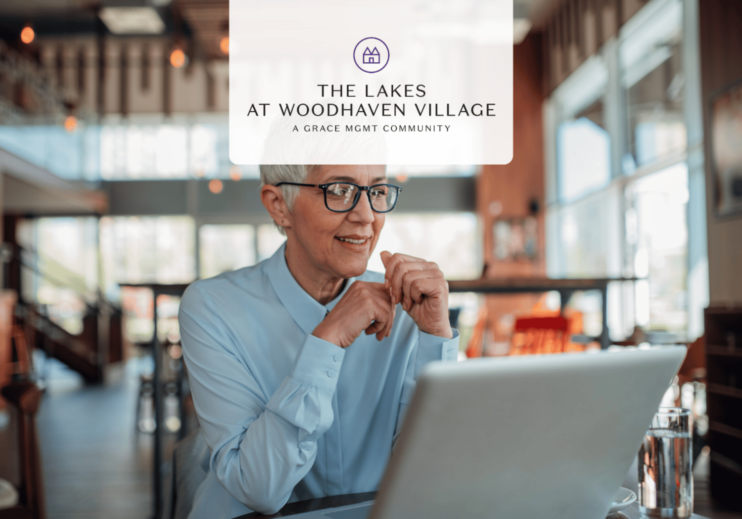 Senior woman with short gray hair and glasses using a laptop in a café; overlay promotes The Lakes at Woodhaven Village community.