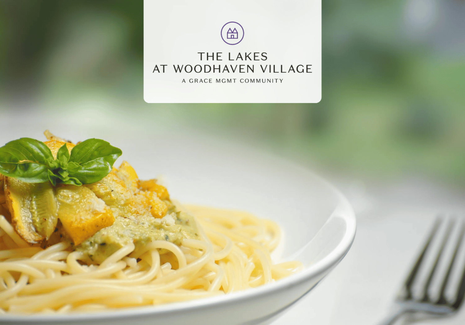 Plate of spaghetti with a green pesto-topped topping and fresh basil on a white plate, against a soft, blurred green background; a branding card in the center reads 'The Lakes at Woodhaven Village'.